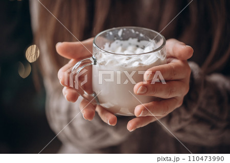 Transparent glass mug of cocoa with marshmallows in female hands. Close-up. New Year's portrait. 110473990
