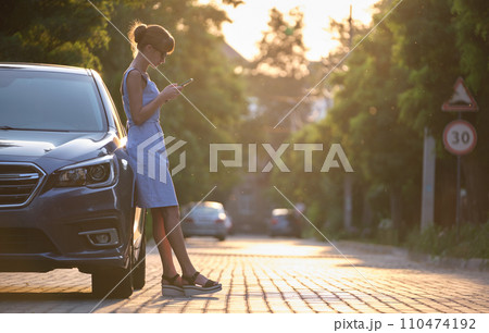 Young female driver standing near her vehicle browsing cellphone on a city street in summer. 110474192