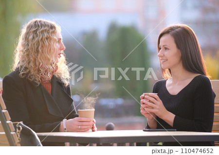 Happy young women friends drinking coffee at city street restaurant during work break. Socializing and friendship concept Happy young women friends drinking coffee at city street restaurant during work break. Socializing and friendship concept 110474652