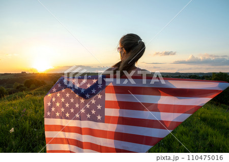 Back view of happy woman with USA national flag standing outdoors at sunset. Positive girl celebrating United States independence day. International day of democracy concept. 110475016