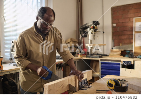 Portrait of African American senior man cutting wood while building furniture in sunlit workshop Portrait of African American senior man cutting wood while building furniture in sunlit workshop 110476882
