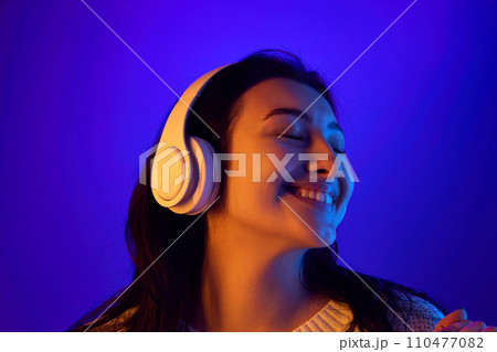 Closed eyes harmony. Close up portrait of young attractive woman listening favorite soundtrack in headphones against blue background in neon. Closed eyes harmony. Close up portrait of young attractive woman listening favorite soundtrack in headphones against blue background in neon. 110477082