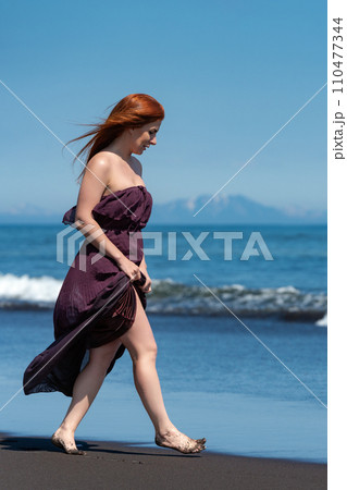 Redhead woman strolling along sandy beach in long dress, with hem raised to her knees, looking down Redhead woman strolling along sandy beach in long dress, with hem raised to her knees, looking down 110477344