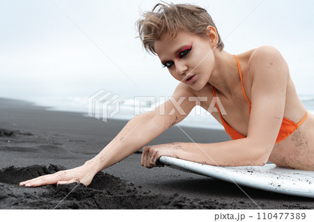 Portrait of blonde surfer woman with bright makeup lying in front of surfboard on sandy beach 110477389
