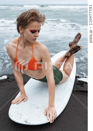Female surfer lies on front of surfboard, arching her back and crossing legs while leaning on hands 110477391