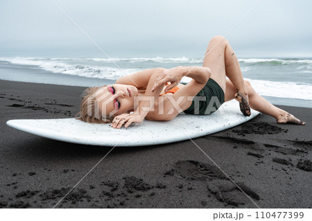 Female surfer lying on white surfboard on black sandy beach. Sports fashion model with closed eyes 110477399