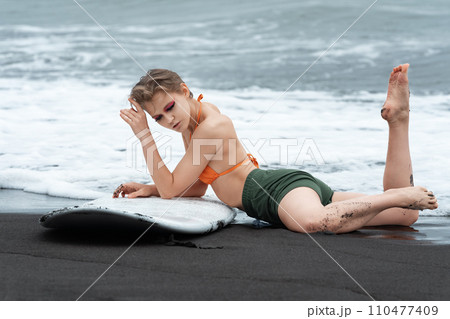 Dreamy sexy surfer is lying on black sandy beach with surfboard, showing off fit and athletic body Dreamy sexy surfer is lying on black sandy beach with surfboard, showing off fit and athletic body 110477409