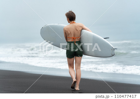 Rear view of unidentified female surfer walking on black sandy beach while carrying white surfboard 110477417