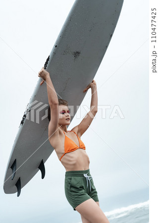 Female surfer carrying surfboard on head during sports training. Woman in bikini top and shorts Female surfer carrying surfboard on head during sports training. Woman in bikini top and shorts 110477425