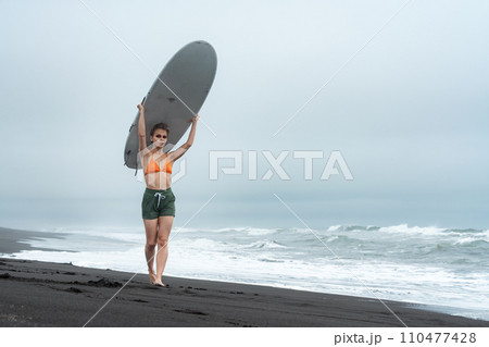 Woman surfer walking on beach and carrying white surfboard on head against background of sea waves Woman surfer walking on beach and carrying white surfboard on head against background of sea waves 110477428