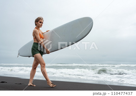 Surfer is walking on sand beach during summer holiday, carrying surfboard on background of sea waves 110477437