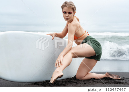 Female surfer exudes confidence as she kneels on black sand beach, holding surfboard with ease 110477447
