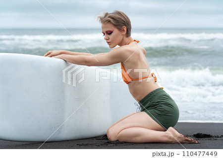 Female surfer, with her eyes closed, is kneeling and holding her surfboard during summer vacation Female surfer, with her eyes closed, is kneeling and holding her surfboard during summer vacation 110477449