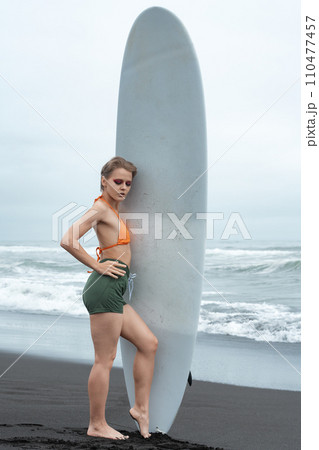 Female surfer stands on beach and holds white surfboard vertical on background of breaking waves 110477457