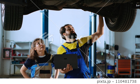 Certified serviceman in garage workspace standing with customer underneath suspended car, looking together for replacement parts. Expert helps woman fix vehicle, using laptop to find components online Certified serviceman in garage workspace standing with customer underneath suspended car, looking together for replacement parts. Expert helps woman fix vehicle, using laptop to find components online 110481738