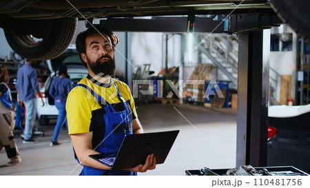 Qualified repairman standing underneath suspended car in garage workspace using laptop to order new parts after finding issues. Repair shop worker looking online for replacing components using device 110481756