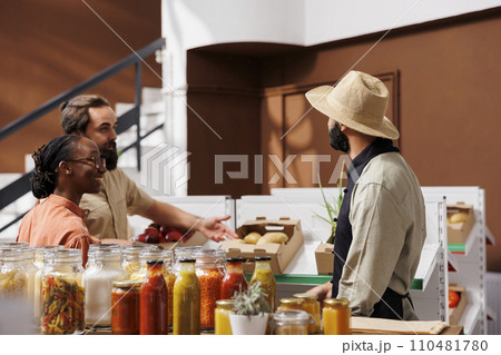 Merchant with a hat showing freshly harvested produce to young husband and wife in local market. Male seller recommending homegrown additives free fruits and veggies to multiracial couple. Merchant with a hat showing freshly harvested produce to young husband and wife in local market. Male seller recommending homegrown additives free fruits and veggies to multiracial couple. 110481780