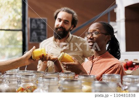 Portrait of multiracial customers actively selecting bio food items at local small business. Boyfriend and girlfriend shopping for natural honey in an eco friendly convenience store. 110481838