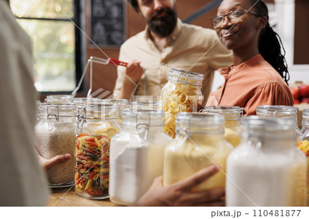 Close-up of african american girlfriend grasping a glass jar full of pasta while standing near caucasian boyfriend with basket. Multiethnic customers shop sustainably for healthy way of life. 110481877