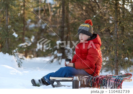 A boy in a red jacket and a knitted hat sits on sled in the winter forest. 110482739