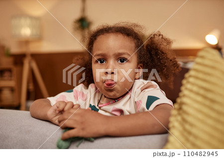 Medium close up shot of African American preschool girl with curly ponytails sticking out her tongue while leaning on couch Medium close up shot of African American preschool girl with curly ponytails sticking out her tongue while leaning on couch 110482945