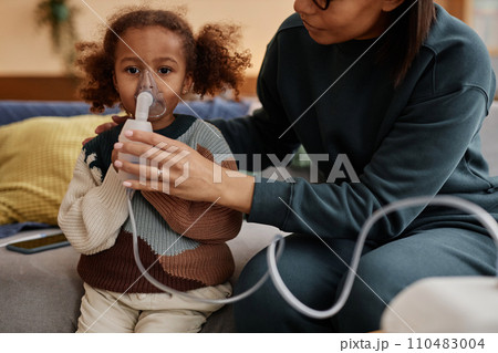 Cropped shot of mother helping big-eyed African American daughter with inhalation holding nebulizer mask at girls face Cropped shot of mother helping big-eyed African American daughter with inhalation holding nebulizer mask at girls face 110483004