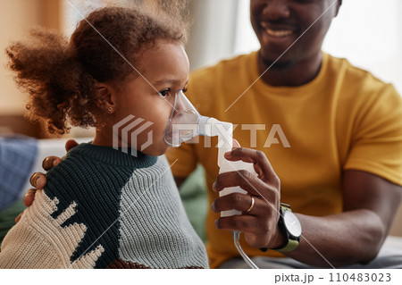 Side view of curly-haired African American preschool girl doing inhalation at home with help of father holding nebulizer mask 110483023