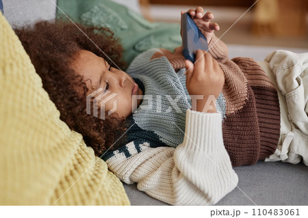 Medium shot of little African American girl in sweater lying on couch at home and playing games using smartphone 110483061