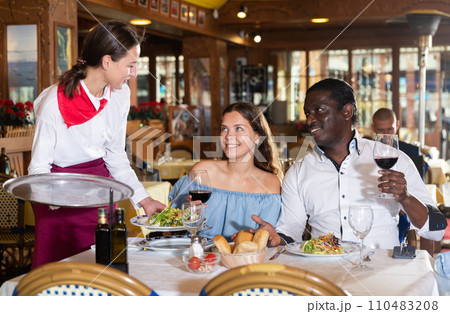 Young waitress bringing dishes to couple at restaurant Young waitress bringing dishes to couple at restaurant 110483208