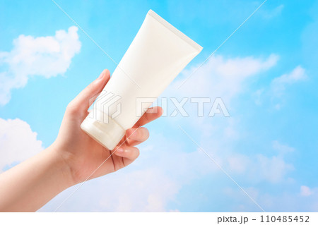 Hand of woman is holding a white plastic tube container for moisturizer or sunscreen on blue sky. Cosmetic bottle for design packaging. Minimal scene for advertising 110485452
