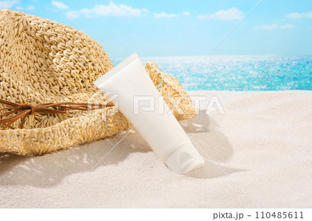Close-up of an unlabeled tube of sunscreen leaning against a straw hat on white sand and blue sea water. Exquisite mockup for advertising. Blank labels for branding. 110485611
