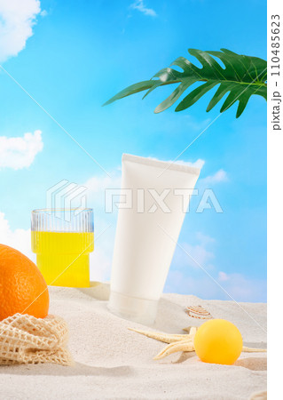 An empty cosmetic tube, orange, juice glass, starfish and tropical leaves are displayed against a background of white sand and blue sky. Cosmetic mockups. An empty cosmetic tube, orange, juice glass, starfish and tropical leaves are displayed against a background of white sand and blue sky. Cosmetic mockups. 110485623