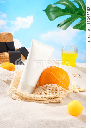 Front view of a white unlabeled cosmetic tube leaning against an orange, in the background is a straw hat and a glass of juice. Tropical leaves. Copy space. Front view of a white unlabeled cosmetic tube leaning against an orange, in the background is a straw hat and a glass of juice. Tropical leaves. Copy space. 110485624