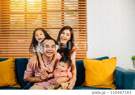 A cheerful family in their comfortable modern house shares a sweet moment on the sofa. Parents daughters and siblings bond laugh and embrace radiating joy and togetherness. A cheerful family in their comfortable modern house shares a sweet moment on the sofa. Parents daughters and siblings bond laugh and embrace radiating joy and togetherness. 110487165
