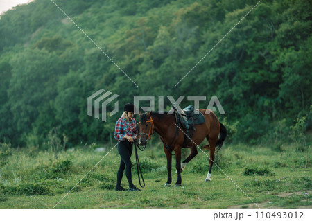 Happy blonde with horse in forest. Woman and a horse walking through the field during the day. Dressed in a plaid shirt and black leggings. Happy blonde with horse in forest. Woman and a horse walking through the field during the day. Dressed in a plaid shirt and black leggings. 110493012