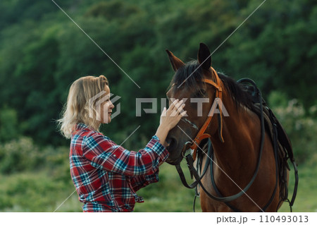 Happy blonde with horse in forest. Woman and a horse walking through the field during the day. Dressed in a plaid shirt and black leggings. Happy blonde with horse in forest. Woman and a horse walking through the field during the day. Dressed in a plaid shirt and black leggings. 110493013