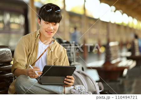 Male traveler using digital tablet while sitting on a bench at the train station Male traveler using digital tablet while sitting on a bench at the train station 110494721