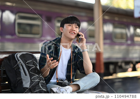 Cheerful young male traveler talking on mobile phone while waiting for the train at the train station Cheerful young male traveler talking on mobile phone while waiting for the train at the train station 110494788