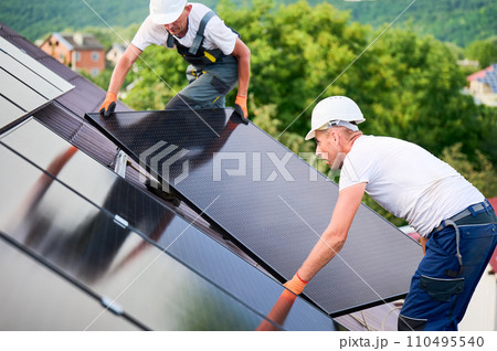 Workers building solar panel system on rooftop of house. Two men installers in helmets installing photovoltaic solar module outdoors. Alternative, green and renewable energy generation concept. 110495540