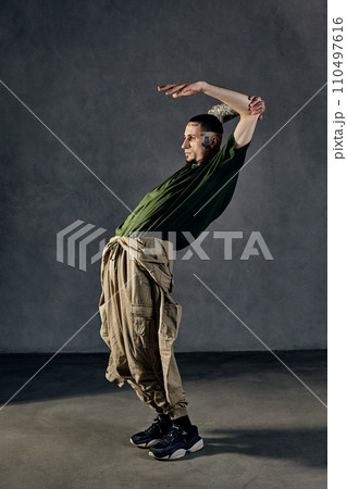 Flexible fellow with tattooed body, earrings, beard. Dressed in khaki t-shirt, jumpsuit, black sneakers. Dancing, gray background. Dancehall, hip-hop Flexible fellow with tattooed body, earrings, beard. Dressed in khaki t-shirt, jumpsuit, black sneakers. Dancing, gray background. Dancehall, hip-hop 110497616