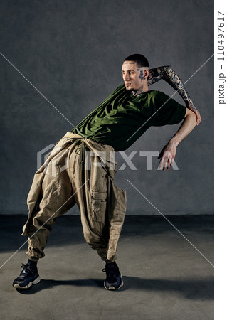 Flexible guy with tattooed body, earrings, beard. Dressed in khaki t-shirt, overalls, black sneakers. Dancing on gray background. Dancehall, hip-hop 110497617