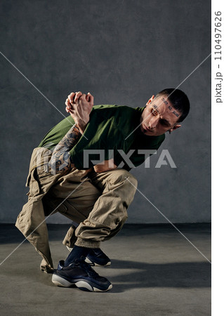 Graceful fellow with tattooed body, earrings, beard. Dressed in khaki t-shirt, jumpsuit, black sneakers. Dancing, gray background. Dancehall, hip-hop 110497626