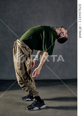 Graceful guy with tattooed body, earrings, beard. Dressed in khaki t-shirt, overalls, black sneakers. Dancing on gray background. Dancehall, hip-hop 110497628