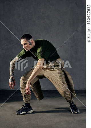 Graceful man with tattooed body, earrings, beard. Dressed in khaki t-shirt, overalls, black sneakers. Dancing on gray background. Dancehall, hip-hop Graceful man with tattooed body, earrings, beard. Dressed in khaki t-shirt, overalls, black sneakers. Dancing on gray background. Dancehall, hip-hop 110497636