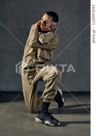 Unusual performer with tattooed body, earrings, beard. Dressed in khaki overalls and black sneakers. Dancing on gray background. Dancehall, hip-hop Unusual performer with tattooed body, earrings, beard. Dressed in khaki overalls and black sneakers. Dancing on gray background. Dancehall, hip-hop 110497643