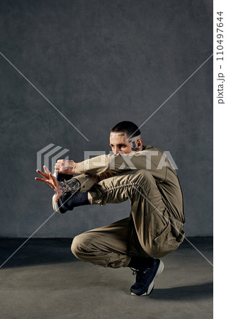 Unusual fellow with tattooed body and face, earrings, beard. Dressed in khaki jumpsuit, black sneakers. Dancing on gray background. Dancehall, hip-hop Unusual fellow with tattooed body and face, earrings, beard. Dressed in khaki jumpsuit, black sneakers. Dancing on gray background. Dancehall, hip-hop 110497644