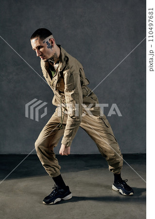 Active fellow with tattooed body and face, earrings, beard. Dressed in khaki jumpsuit, black sneakers. Dancing on gray background. Dancehall, hip-hop 110497681