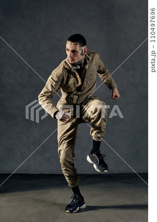 Stately performer with tattooed body, earrings, beard. Dressed in khaki overalls and black sneakers. Dancing on gray background. Dancehall, hip-hop 110497696