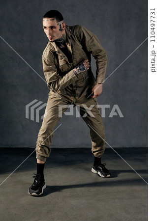 Modern performer with tattooed body, earrings, beard. Dressed in khaki overalls and black sneakers. Dancing on gray background. Dancehall, hip-hop Modern performer with tattooed body, earrings, beard. Dressed in khaki overalls and black sneakers. Dancing on gray background. Dancehall, hip-hop 110497731