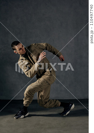Graceful male with tattooed body and face, earrings, beard. Dressed in khaki jumpsuit, black sneakers. Dancing on gray background. Dancehall, hip-hop Graceful male with tattooed body and face, earrings, beard. Dressed in khaki jumpsuit, black sneakers. Dancing on gray background. Dancehall, hip-hop 110497740